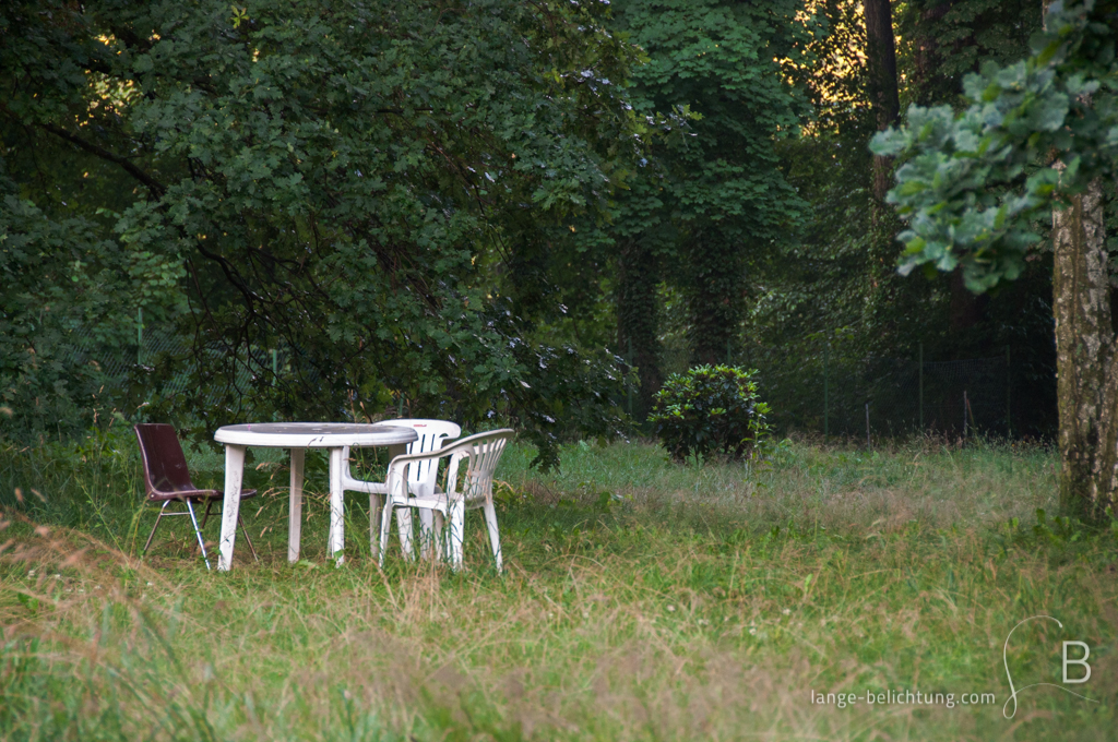 Zwei weiße und ein dunkelroter Stuhl stehen um einen weißen Gartentisch mitten auf der Wiese unter einem Baum. Es wirkt so, als würde man auf jemanden warten.