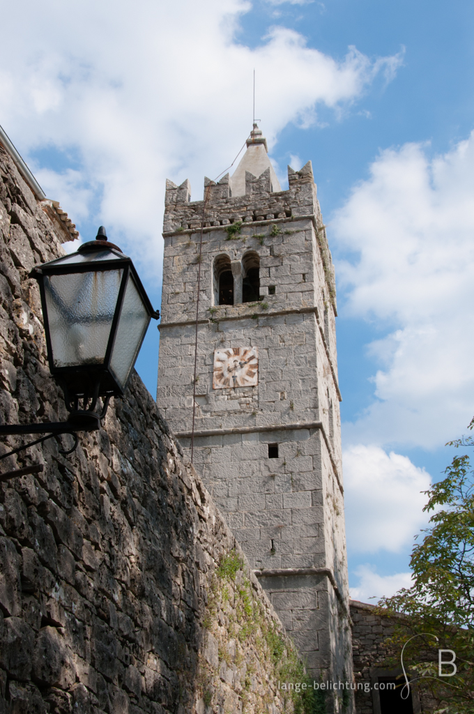 Der Kirchturm von Hum in Kroatien mit alter verrosteter Uhr. Im Vordergrund ist eine als Laterne an der Stadtmauer zu sehen.
