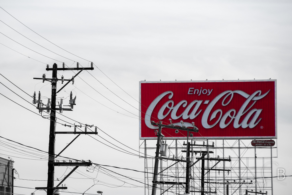 Eine Coca Cola Werbetafel hinter einer Vielzahl von Strommasten und Überlandleitungen an einer Straße.